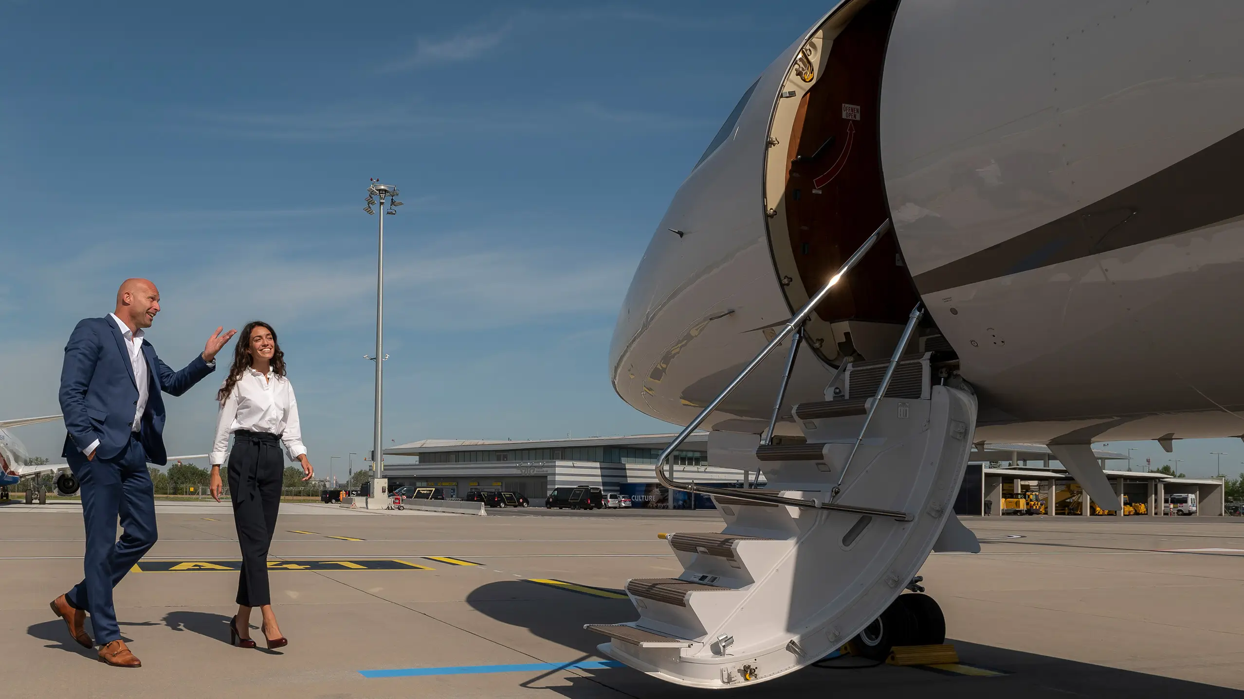 Two professionally dressed people walking toward a private jet on the tarmac, engaged in conversation and smiling. The open cabin stairs and clear skies suggest luxury, ease, and a welcoming charter brokerage experience.