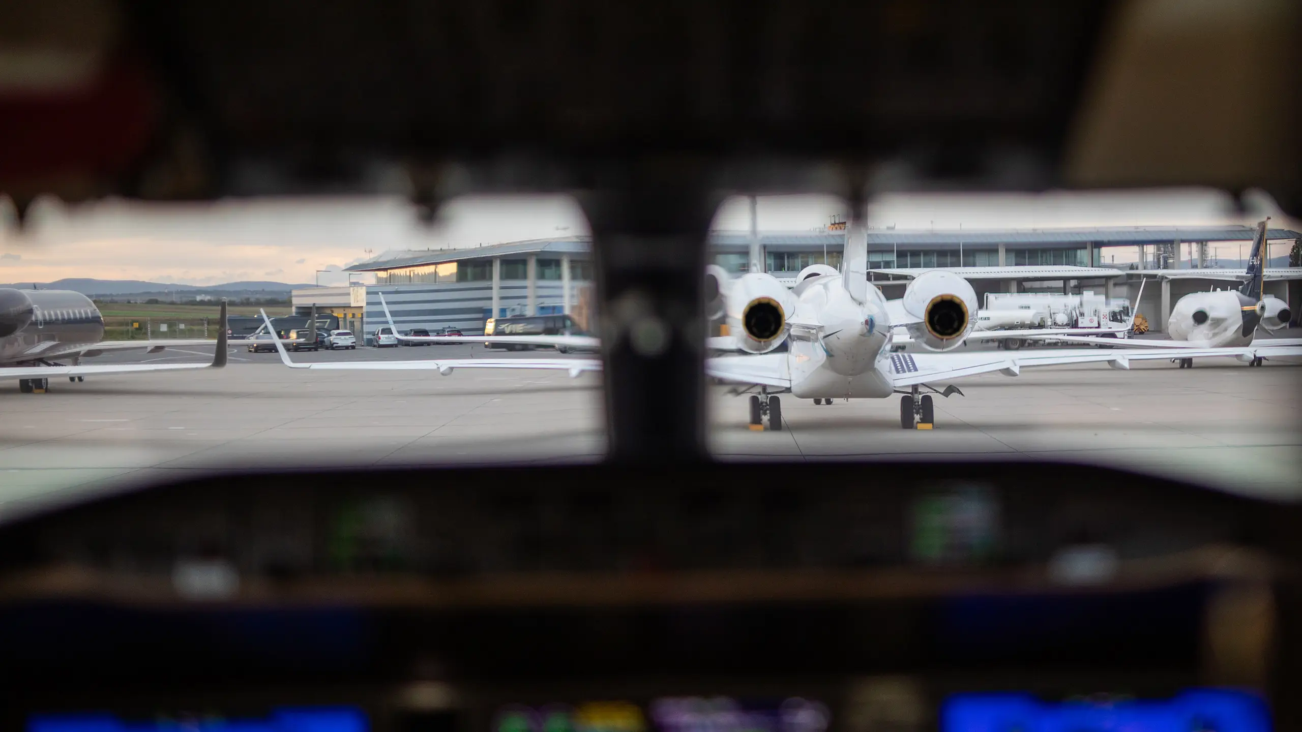 View from inside a cockpit looking out onto an airport tarmac with multiple private jets parked. The central focus is a white jet with engines visible, facing the cockpit, with a modern terminal building and service vehicles in the background.